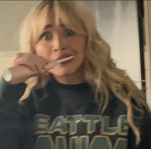 A grainy, slightly blurry selfie of a woman with blonde, wavy hair and bangs. She is wearing a dark navy or black sweatshirt with "SEATTLE" written across the chest in large, blocky green letters. She has an wide-eyed, humorous expression while holding a pink electric toothbrush to her mouth as if brushing her teeth.