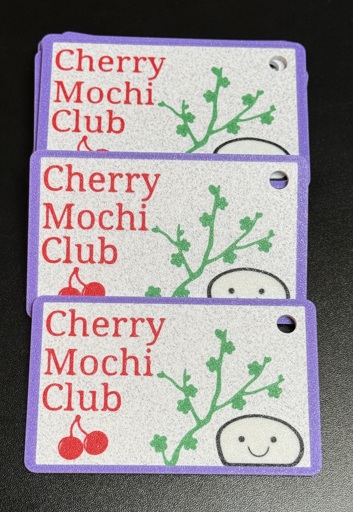 A small stack of 3d printed cards, with the text Cherry Mochi Club on each of them, along with a styalized cherry, cherry blossom branch, and a piece of mochi with a smiling face on it.